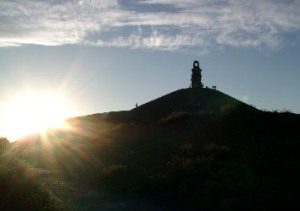 Die Halde Rheinelbe mit der Skulptur Himmelstreppe hebt sich tiefschwarz im Gegenlicht vom blauen Himmel ab. Die untergehende Sonne strahlt hinter dem Haldenhügel hervor.